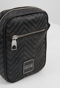 Borsa sintetica nera con motivo ripetuto del logo 'Versace Jeans Couture', chiusura con zip e accento quadrato del logo sul davanti.