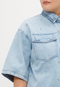 Lys blå denimskjorte med korte ermer, to brystlommer og sølvnagler som dekorasjon på forsiden. Klassisk kragedesign.