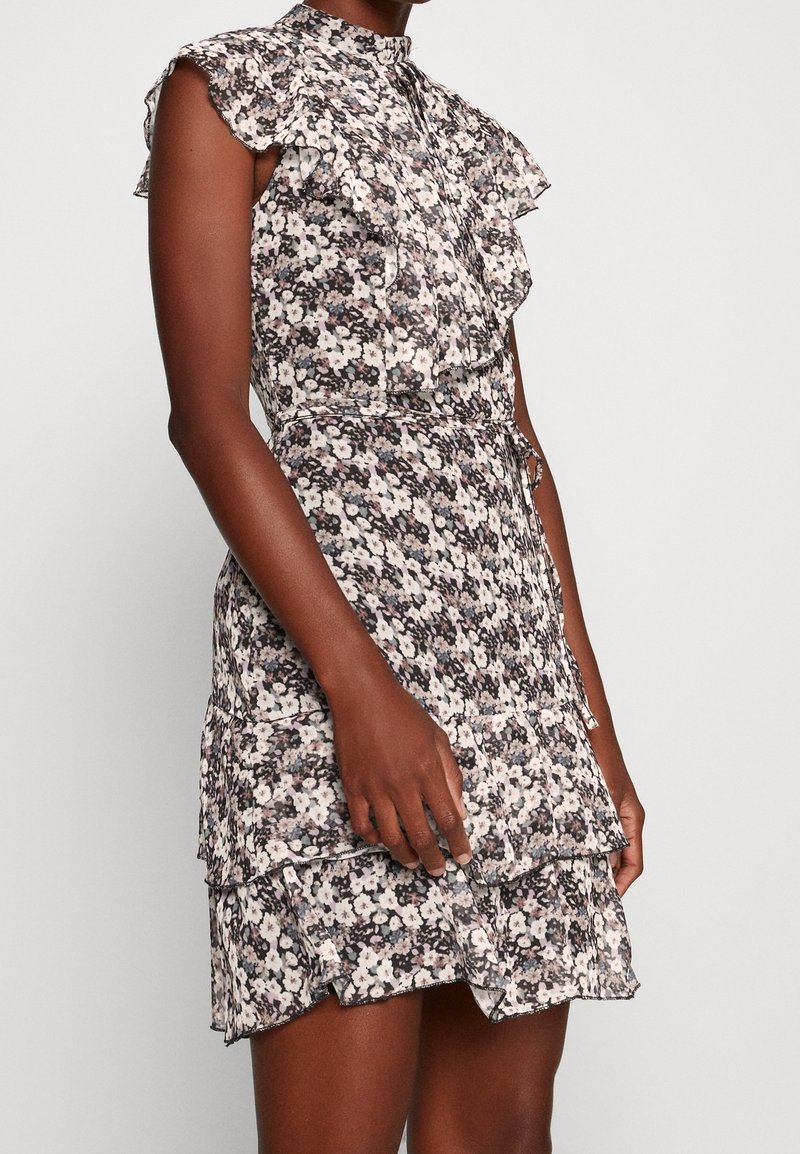 Robe sans manches à fleurs avec des volants et un col montant, portée par une personne à la peau foncée sur un fond clair uni.