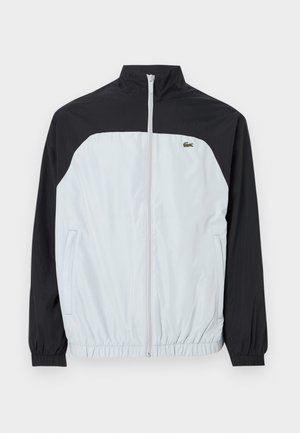 Biela a čierna vetrovka s vysokým golierom, zipsom vpredu, elastickými manžetami a malým zeleným logom na hrudi.