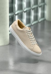 Beige semsket sneaker med en hvit gummisåle, med flate lisser, rund tå og subtile sømdetaljer.