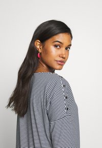 Camisa de mangas longas listrada em azul marinho e branco com botões no ombro. Modelo com brincos de argola cor-de-rosa e cabelo longo e liso.