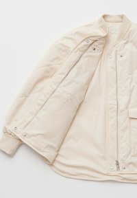 Veste matelassée beige clair avec poignets côtelés, fermeture éclair argentée et boutons-pression, partiellement dézippée montrant une doublure intérieure lisse et une poche intérieure.