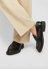 Mocassins de couro preto com uma costura elevada e em relevo, bico redondo e sola baixa em plataforma preta, combinados com calças largas bege.