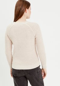 Pull en laine beige avec une texture côtelée, col rond, manches raglan et ourlet accentué. Associé à un jean gris foncé.