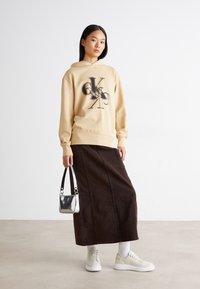 Haut à capuche beige avec un design graphique noir, jupe maxi en denim marron, baskets blanches et un petit sac bandoulière en argent. Textures lisses partout.
