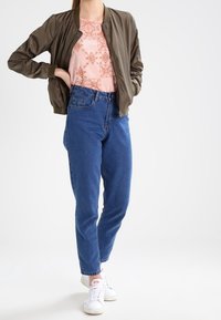 Olivově zelená bomber bunda, světle růžový top s květinovým vzorem, modré džíny s vysokým pasem, bílé tenisky, volný střih, hladká textura.