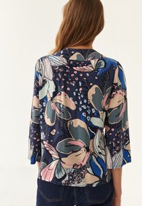 Blusa floral con mangas tres cuartos, fondo azul marino, grandes patrones florales multicolores y textura sedosa. Presenta un escote redondo.