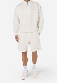Sweat-shirt blanc à col zippé avec épaules tombantes, associé à un short léger assorti. Tissu doux et branding discret sur les deux pièces.