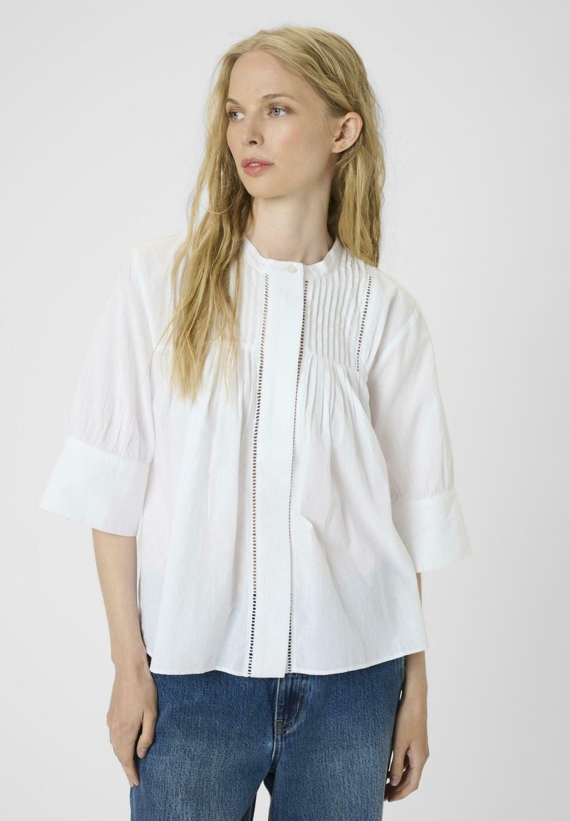 White cotton blouse with a round neck, pleated front, and button closure. Features short sleeves and a relaxed fit, paired with blue jeans.