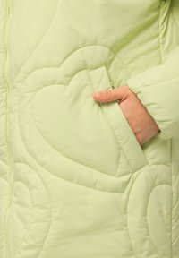 Abrigo acolchado verde claro con costuras en forma de corazón y un bolsillo lateral con una mano visible en su interior. Tejido suave y texturizado.