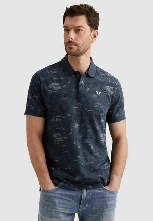 Man met kort haar die een donkerblauw polohemd met patroon en kraag draagt en een lichtblauwe spijkerbroek, staat met handen in de zakken.
