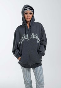 Dark gray zip-up hoodie features a large, bold, white graphic across the chest. Material appears soft, with a relaxed fit and hood.