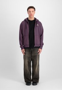 Fialová mikina na zips s prednými vreckami, čierne tričko pod ňou, voľné tmavé denimové nohavice a čierne topánky. Minimalistický dizajn, žiadne vzory.