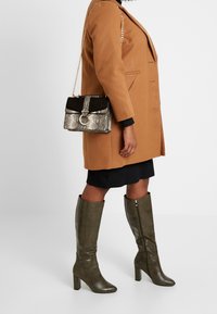 Hnědý kabát v kombinaci s černou a stříbrnou kabelkou ve vzoru hadí kůže. Olivově zelené kozačky po kolena se strukturovaným vzorem a blokovými podpatky.