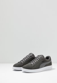 Sneakers in suede grigio con profilo basso, suole in gomma bianca piatta e lacci grigi scuri. Presentano un logo discreto sul lato e sulla linguetta.