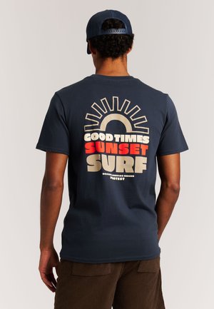 Marineblauw katoenen T-shirt met een grafisch ontwerp van een zon en de tekst "GOOD TIMES SUNSET SURF" in gedurfde, contrasterende kleuren.