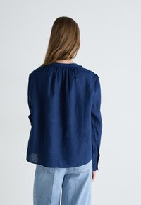 Camicia in lino blu navy con scollo arricciato e maniche lunghe, indossata su jeans di denim azzurro. Texture liscia, design informale.