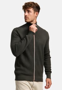 Mørkegrøn strikket sweater med høj krave, med en frontlynlås og en kontrasterende lys orange træk. Ribbet manchetter og kant.