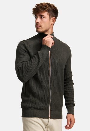 Suéter de punto verde oscuro con un cuello alto, que presenta un cierre frontal con un tirador de color naranja claro en contraste. Puños y dobladillo acanalados.