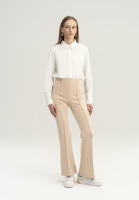 Camisa blanca de botones y pantalones acampanados beige de cintura alta. Textura de tela suave. Zapatillas blancas con un sutil acento de color en el costado.