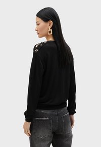 Svart sweatshirt med långa ärmar, med skärningar på axlarna som är fästa med bronserade öglor. Matchas med slitna grå jeans.