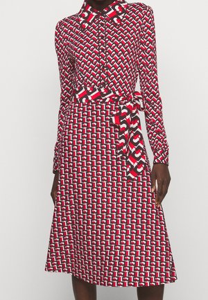 Femme portant une robe à manches longues boutonnée avec un motif géométrique rouge, noir et blanc et une ceinture assortie à la taille.
