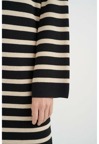 Svart och beige randig tröja med ribbad struktur, som har långa ärmar och rak fåll, där en hand vilar delvis på tyget.