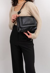 Borsa a tracolla in pelle nera con patta frontale, dettaglio della tracolla annodata e logo impresso. Texture liscia con forma strutturata.