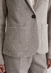 Tan textured blazer made of wool blend with two front pockets and a single button closure. Paired with matching trousers.