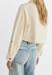 Béžová zkrácená bunda s texturovanou látkou, zapínáním na knoflíky na rukávech a volným střihem, doplněná světle modrými džíny s vysokým pasem.