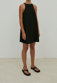 Vestido negro sin mangas con una silueta fluida, hombros descubiertos, acompañado de sandalias planas negras con suelas gruesas y correas.