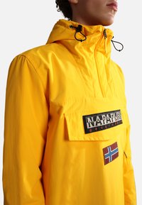 Yellow waterproof jacket with a hood, featuring a front pocket, logo patch, and flag detail. Smooth texture and lightweight design.