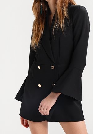 Mujer vestida con un vestido blazer negro de doble botonadura, mangas acampanadas y botones dorados, de pie frente a un fondo blanco.