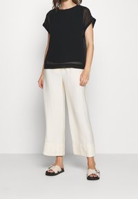 Schwarzes, transparentes Kurzarm-Top, kombiniert mit cremefarbenen, weitem Hosen, die eine glatte Textur und eine gerade Silhouette aufweisen, ergänzt durch leichte Sandalen.