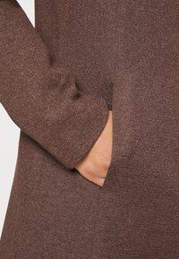Brown, textured fabric with a close-up of a side pocket. Soft, knitted material; sleeve hem is slightly rounded.