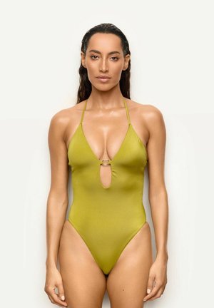 Femme aux longs cheveux foncés portant un maillot de bain une pièce vert à col halter, debout devant un fond clair uni.