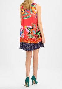 Květinové šaty bez rukávů s červeným vrškem zdobeným živými vzory, přecházející do námořnicky modrého spodku se zlatým vířivým motivem. Zelené vysoké podpatky.