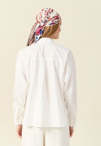 Camicia di cotone bianca con yoke posteriore, maniche lunghe e polsini con bottoni. Il modello indossa un foulard stampato, esposto su uno sfondo neutro.