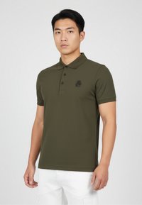 Olivgrön polo skjorta gjord av mjukt tyg, med en placket med tre knappar och en subtil logga på vänster bröst. Korta ärmar.