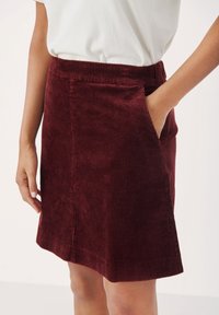 Falda de pana burdeos con una cintura ajustada, bolsillos laterales y textura de canalé vertical; hasta la rodilla con un dobladillo ligeramente acampanado.