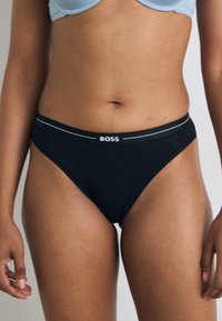 Svart bomullstruse med en kontrasterende grå midjebånd med "BOSS"-logo. Enkel design, jevn tekstur og standard snitt.