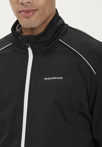 Sort sportsjakke med høj krave. Har hvid lynlås og piping samt "ENDURANCE" logo på forsiden. Materialet ser glat og fleksibelt ud.