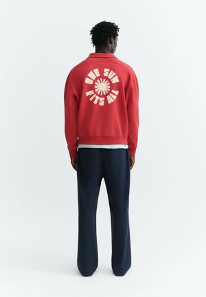 Homme vu de dos portant une veste rouge avec "ONE SUN FITS ALL" et un motif de soleil dans le dos, assortie à un pantalon bleu foncé.