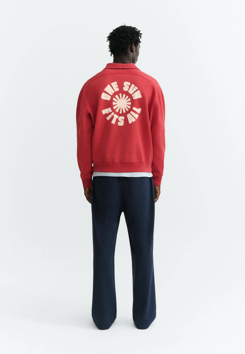 Homme vu de dos portant une veste rouge avec "ONE SUN FITS ALL" et un motif de soleil dans le dos, assortie à un pantalon bleu foncé.