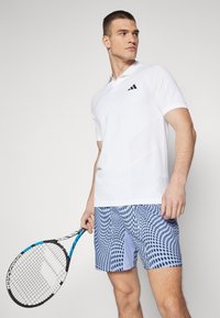 Camisa polo blanca de manga corta, con un logo negro. Combinada con pantalones cortos de patrón azul claro con acentos en azul oscuro, sosteniendo una raqueta de tenis.