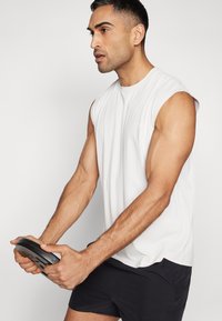 Weißes ärmelloses Sportshirt aus strukturiertem Stoff, dunkle Shorts und eine Gewichtsscheibe in den Händen gehalten. Sauberes, minimalistisches Design.