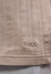 Κοντινή λήψη μπεζ υφάσματος με υφασμένα κάθετα σχέδια ψαροκόκαλο και πλεξούδα και το κεντημένο κείμενο "HUGO" κοντά στο κάτω άκρο.