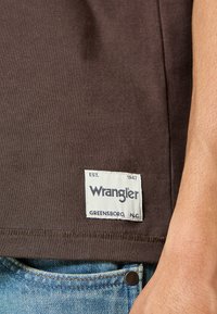 T-shirt in cotone marrone con un'etichetta bianca riportante "Wrangler EST. 1947" e "Greensboro, N.C." ricamati sull'orlo inferiore.