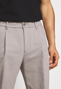 Lichtgrijze broek met plooien, een knoopsluiting en riemlussen, gemaakt van gladde stof, getoond naast een zwart overhemd.